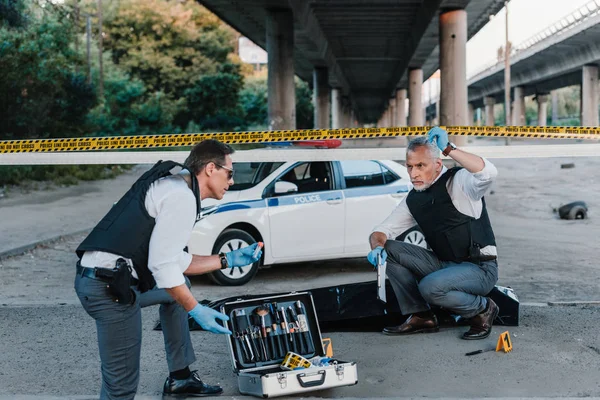 Polizist mit Klemmbrett spricht mit Kollegin mit Sonnenbrille nahe Leiche im Leichensack am Tatort — Stockfoto