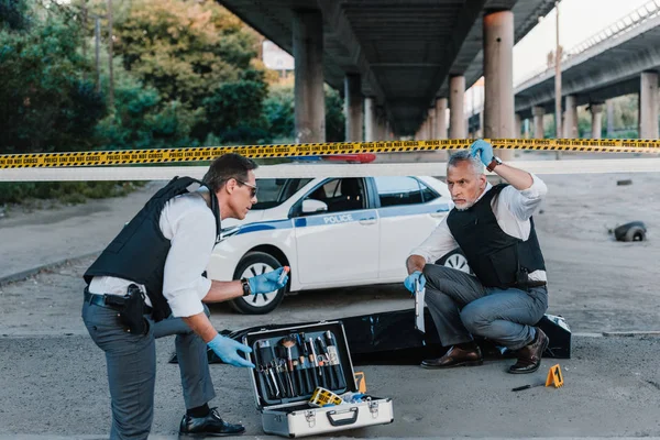 Male police offier with clipboard talking to colleague in sunglasses near corpse in body bag at crime scene — Stock Photo