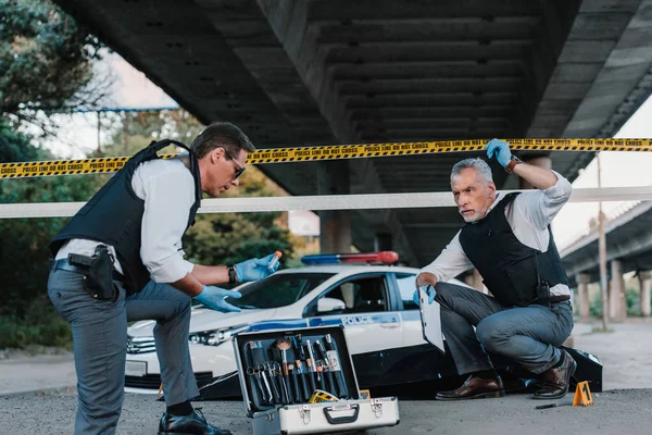 Middle aged policeman holding police line above head and talking to colleague in sunglasses standing near case with investigation tools at crime scene — Stock Photo