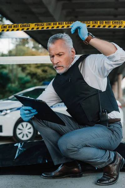 Policier masculin en gants de latex tenant la ligne de police au-dessus de la tête et regardant le presse-papiers près du cadavre dans un sac mortuaire sur les lieux du crime — Photo de stock