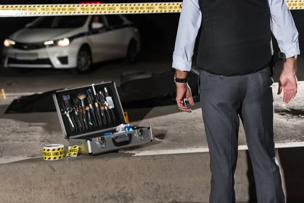 Imagen recortada del policía de pie cerca de la línea transversal y el caso con herramientas de investigación cerca del cadáver en la bolsa de cadáveres en la escena del crimen - foto de stock
