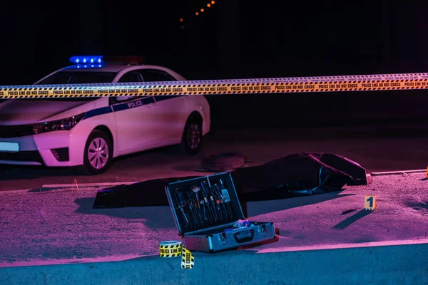 Toned picture of crime scene with police car, case with investigation tools, cross line and corpse in body bag — Stock Photo