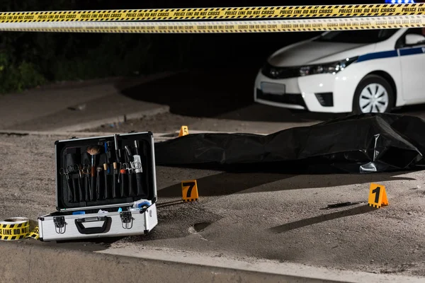 Caso con herramientas de investigación, coche, línea de policía y cadáver en bolsa de cuerpo en la escena del crimen - foto de stock
