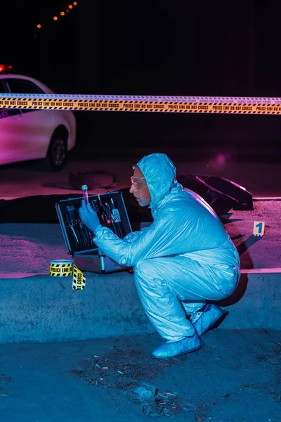 Seitenansicht des fokussierten männlichen Kriminologen in Schutzanzug und Latexhandschuhen, der am Tatort mit Leiche Beweise sammelt — Stockfoto