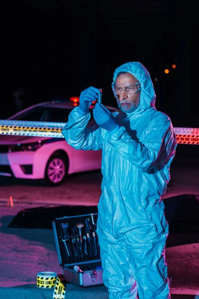 Criminologue masculin concentré en combinaison de protection et gants en latex recueillant des preuves près de la ligne transversale sur les lieux du crime — Photo de stock