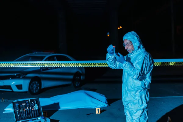 Criminólogo varón maduro en traje protector y guantes de látex recogiendo pruebas en la escena del crimen con el cadáver - foto de stock