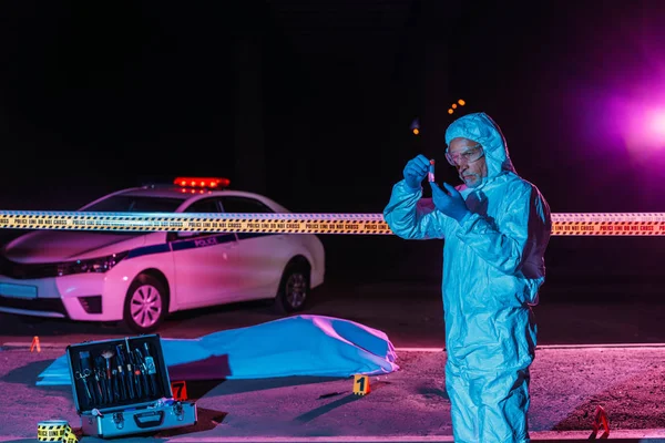 Middle aged male criminologist in protective suit and latex gloves collecting evidence at crime scene with corpse — Stock Photo