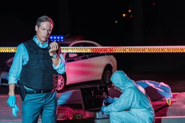 Mature male police officer talking on radio set while criminologist collecting evidence at crime scene with corpse — Stock Photo