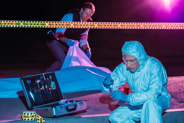 Criminólogo masculino de mediana edad recogiendo pruebas mientras el policía cubre el cadáver en la escena del crimen - foto de stock