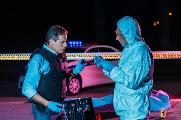 Male criminologist in protecitve suit showing evidence to mature policeman at crime scene with corpse in body bag — Stock Photo