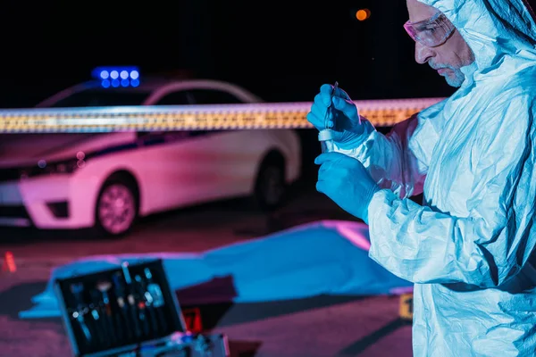 Focused male criminologist in protective suit and latex gloves putting evidence into flask by tweezers at crime scene with corpse — Stock Photo