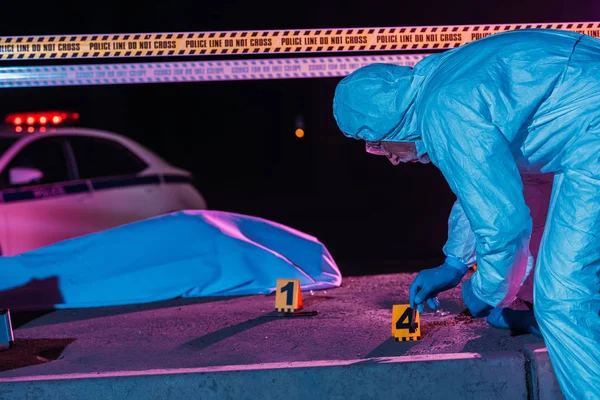 Criminólogo masculino de mediana edad en traje protector y guantes de látex recogiendo evidencia en la escena del crimen con cadáver - foto de stock