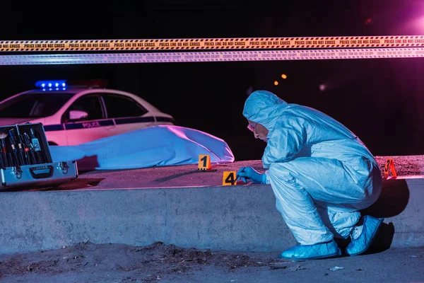 Criminólogo masculino en traje de protección y guantes de látex recogiendo pruebas en la escena del crimen con el cadáver - foto de stock