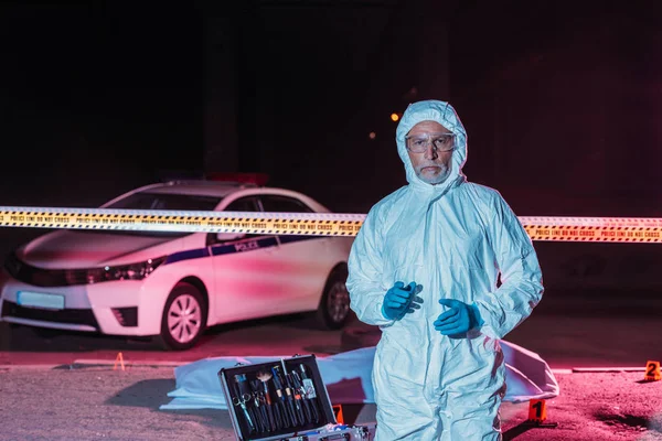 Mature male criminologist in protective suit and mask looking at camera near crime scene with corpse in body bag — Stock Photo