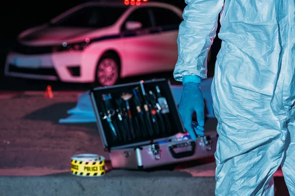 Cropped image of male male criminologist in protective suit standing near case with investigation tools at crime scene — Stock Photo