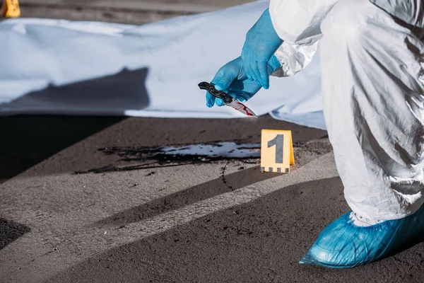 Partial view of criminologist in latex gloves and protective suit holding knife above blood on ground near corpse in body bag at crime scene — Stock Photo