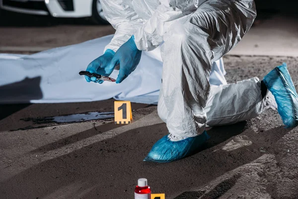 Imagem cortada de criminologista em luvas de látex e traje protetor segurando faca acima do sangue no chão perto do cadáver em saco de corpo na cena do crime — Fotografia de Stock