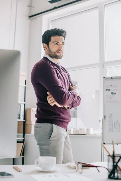 Beau designer en pull bordeaux debout avec les bras croisés et regardant loin dans le bureau — Photo de stock