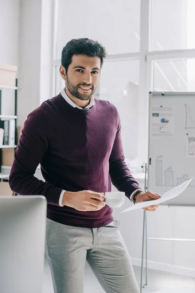 Guapo hombre de negocios en jersey de color burdeos sosteniendo taza de café y documentos en la oficina - foto de stock