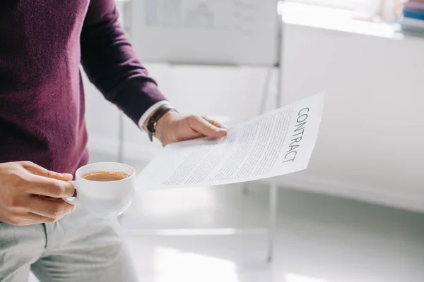 Immagine ritagliata di designer in possesso di tazza di caffè e contratto in ufficio — Foto stock