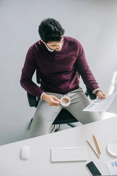 Vista de ángulo alto del diseñador guapo en suéter de color burdeos que sostiene la taza de café y la lectura de documentos en la oficina - foto de stock