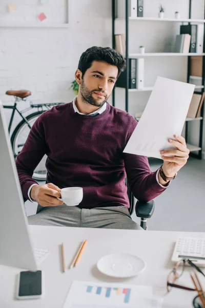 Handsome businessman in burgundy sweater holding cup of coffee and reading documents in office — Stock Photo