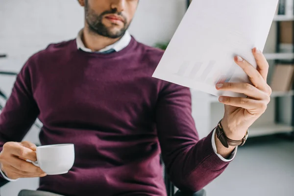 Abgeschnittenes Bild des Designers mit Borsten, die eine Tasse Kaffee halten und Dokumente im Büro lesen — Stockfoto