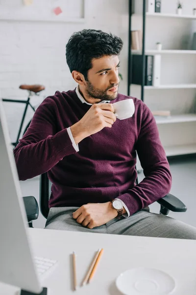 Handsome businessman in burgundy sweater drinking coffee in office and looking away — Stock Photo