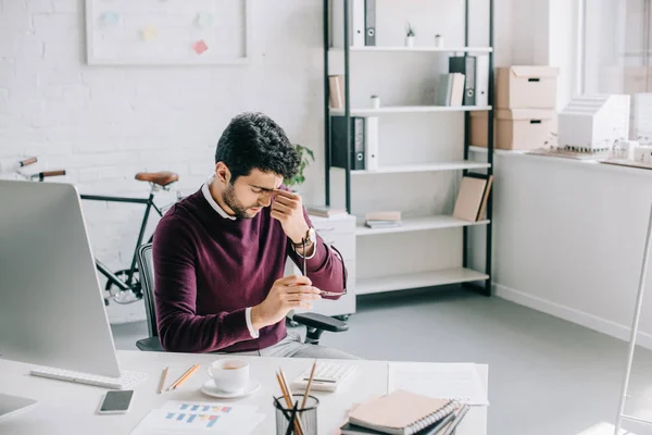 Müder Geschäftsmann im weinroten Pullover rührt Nasenbrücke an und hält Brille im Büro — Stockfoto