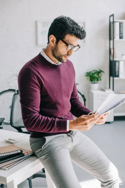 Guapo diseñador en jersey de color burdeos apoyado en la mesa y la lectura de documentos en la oficina - foto de stock