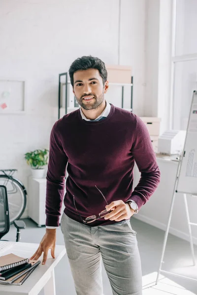 Sorrindo designer bonito em camisola borgonha segurando óculos e olhando para a câmera no escritório — Fotografia de Stock