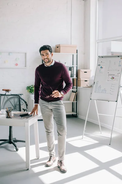 Smiling handsome businessman in burgundy sweater holding glasses and looking at camera in office — Stock Photo