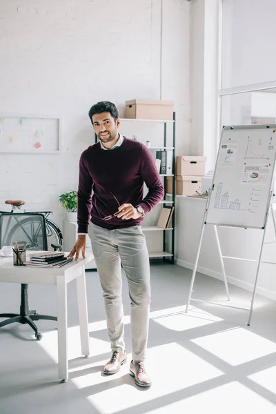 Homme d'affaires beau et souriant en pull bordeaux tenant des lunettes et regardant la caméra dans le bureau — Photo de stock