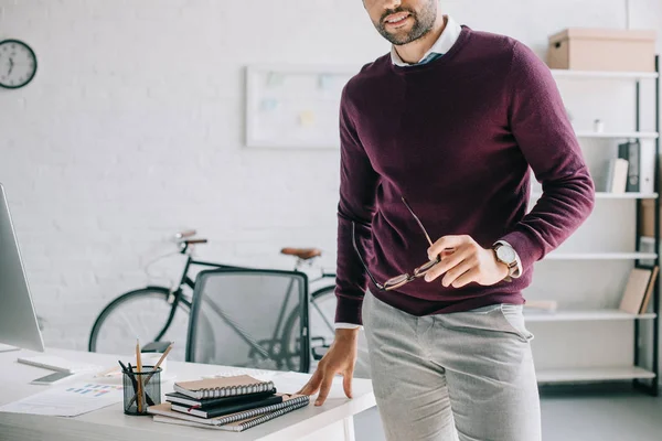 Обрезанный образ улыбающегося бизнесмена в бордовом свитере, держащего очки в офисе — стоковое фото