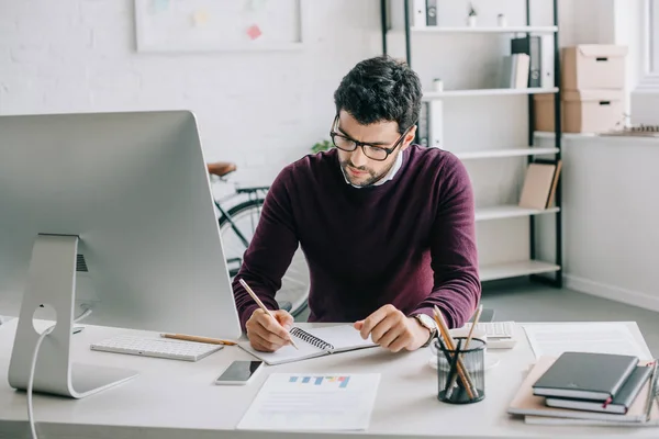 Handsome designer in burgundy sweater writing something to notebook in office — Stock Photo