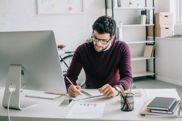 Schöner Designer im weinroten Pullover, der im Büro etwas zum Notizbuch schreibt — Stockfoto