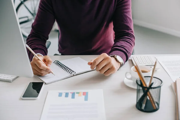 Cropped image of designer in burgundy sweater writing something to notebook in office — Stock Photo