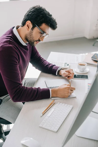 Side view of handsome designer in burgundy sweater making sketch in notebook in office — Stock Photo