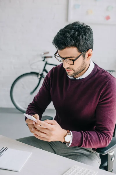 Красивий дизайнер в бордовому светрі, використовуючи смартфон в офісі — стокове фото