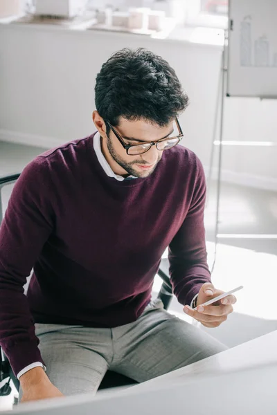 Hombre de negocios guapo en jersey de color burdeos con teléfono inteligente en la oficina - foto de stock