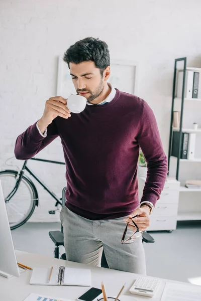 Handsome designer in burgundy sweater drinking coffee and holding glasses in office — Stock Photo