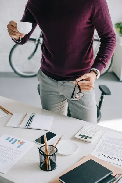 Обрізане зображення дизайнера в бордовому светрі, що тримає чашку кави та окулярів в офісі — стокове фото