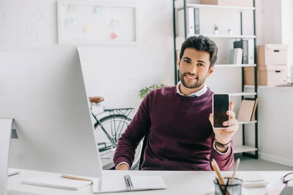 Bel designer sorridente in maglione bordeaux che mostra smartphone con schermo vuoto in ufficio — Foto stock