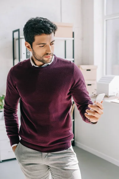 Handsome designer in burgundy sweater looking at smartphone in office — Stock Photo