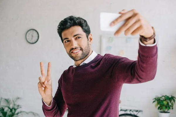 Hombre de negocios sonriente en jersey borgoña tomando selfie con teléfono inteligente y mostrando dos dedos en la oficina - foto de stock