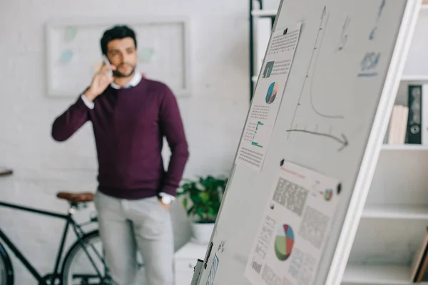 Handsome designer in burgundy sweater talking by smartphone in office with flipchart on foreground — Stock Photo