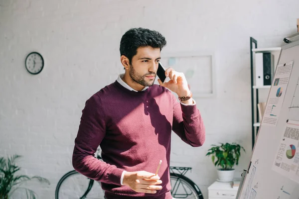 Schöner Geschäftsmann im weinroten Pullover, der im Büro per Smartphone spricht und Flipchart betrachtet — Stockfoto