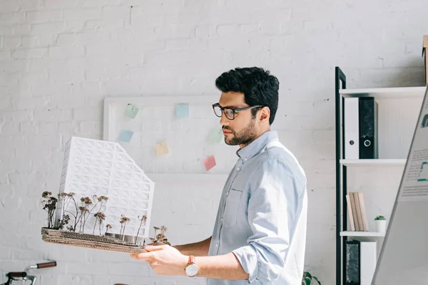 Seitenansicht eines gutaussehenden Architekten mit Brille, der ein Architekturmodell im Büro hält — Stockfoto