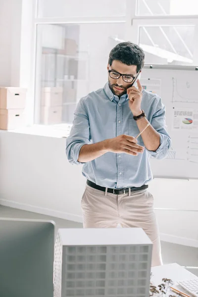 Gutaussehender Architekt spricht per Smartphone und betrachtet Architekturmodell im Büro — Stockfoto