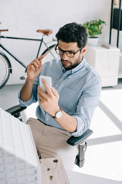 Arquiteto sério usando smartphone perto do modelo de arquitetura no escritório — Fotografia de Stock